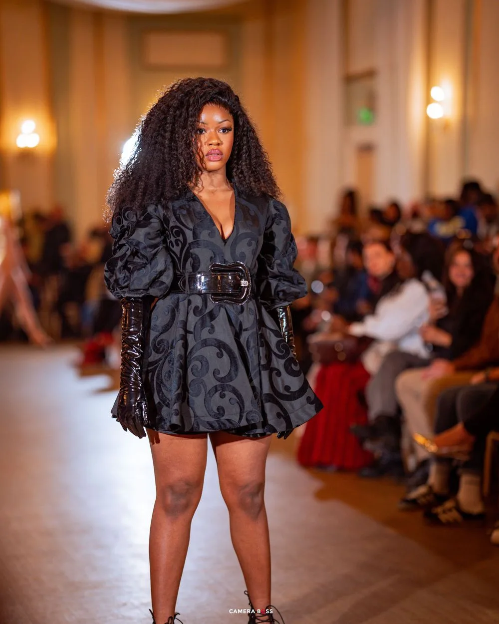 A model walks down a fashion runway wearing a black patterned mini dress with voluminous sleeves and long leather gloves.