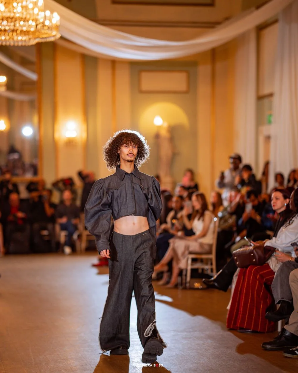 Model showcases gray crop top and wide-leg pants ensemble on runway during fashion show in elegant ballroom setting.