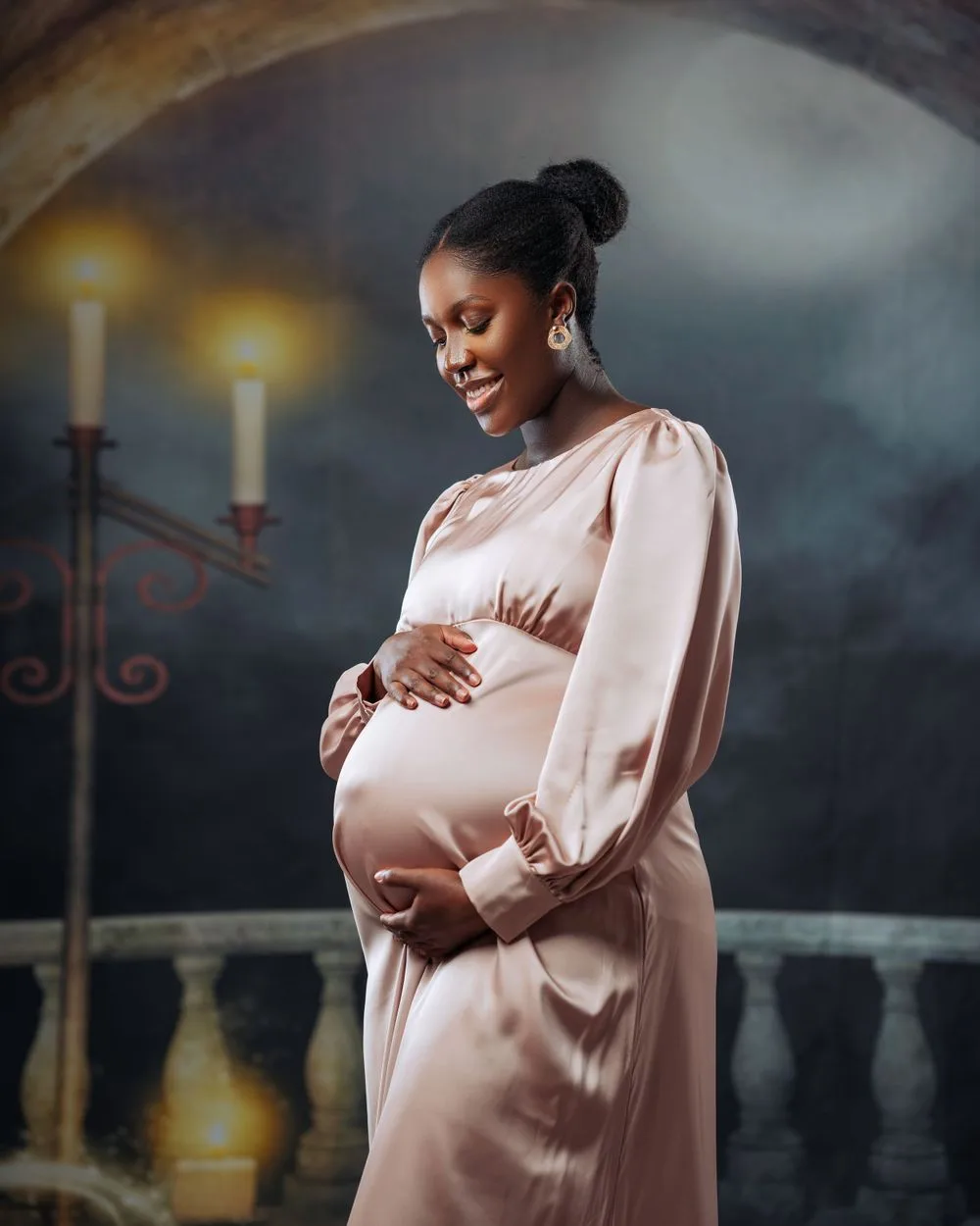 A pregnant woman in a blush pink dress stands elegantly against a moody background with lit candles in the scene.