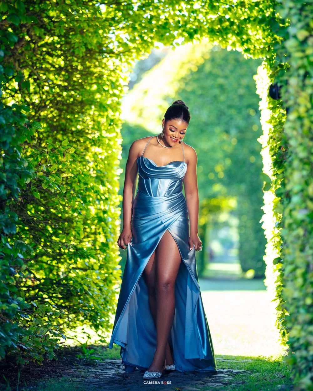 Elegant long blue satin gown with high slit photographed in lush green garden path with sunlit backdrop.