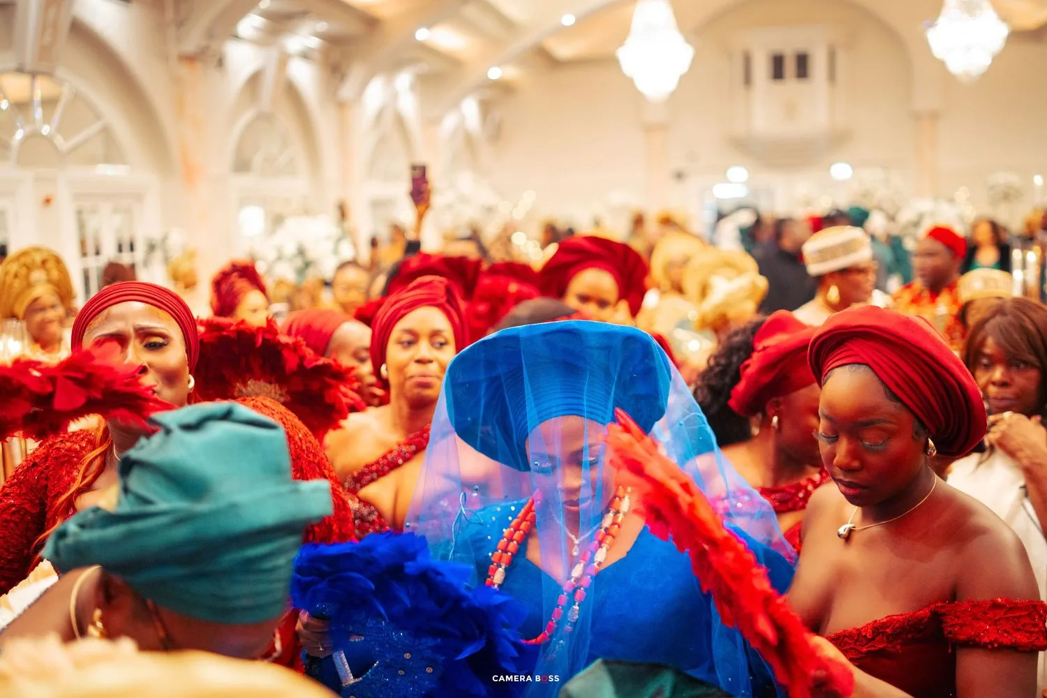 A crowd of people wearing vibrant red and blue outfits and hats gather in an ornately decorated hall.