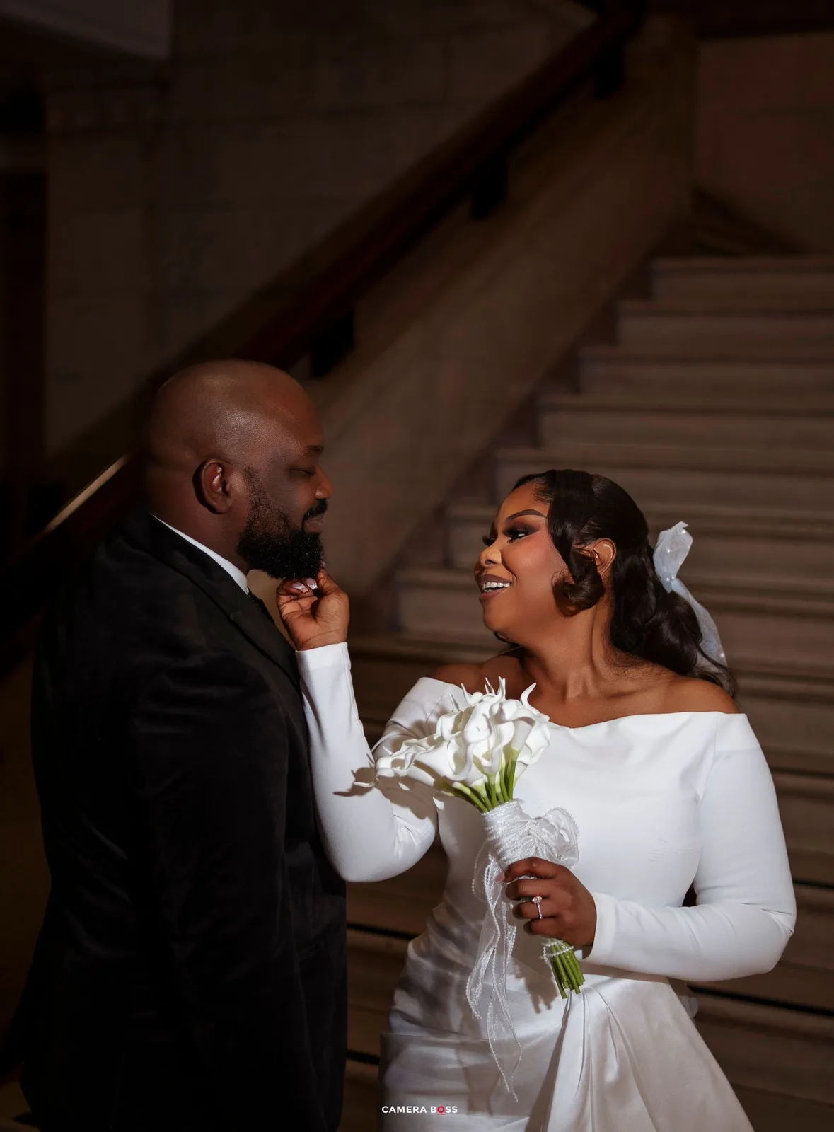 A bride in a white off-shoulder gown holds calla lilies while gazing at her groom in a black tuxedo on a staircase.