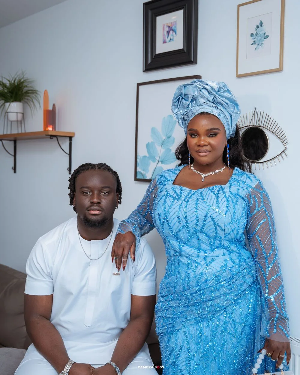 A person in a white t-shirt seated next to someone wearing a blue lace dress and matching headwrap in a room with wall art.