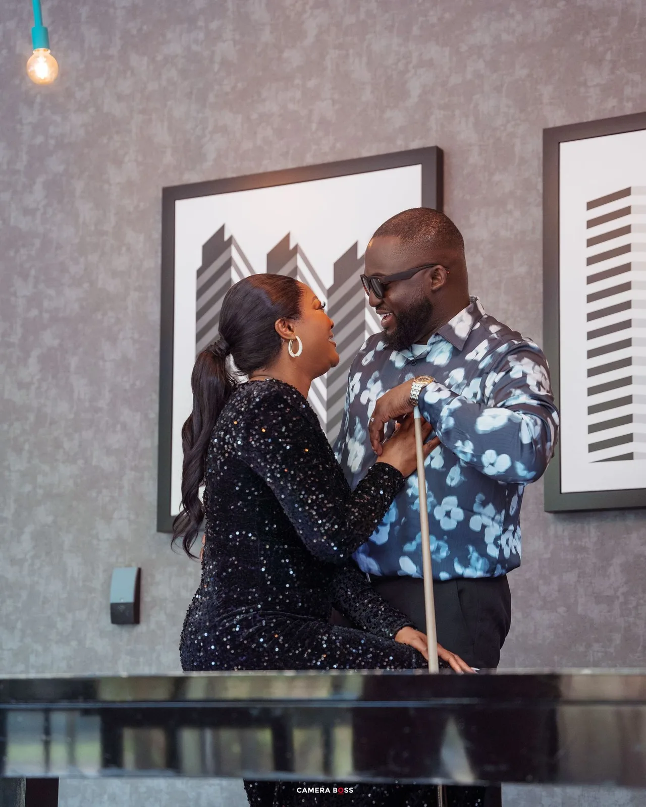 Couple in sparkly black and patterned blue outfits share an intimate moment against grey wall with artwork.