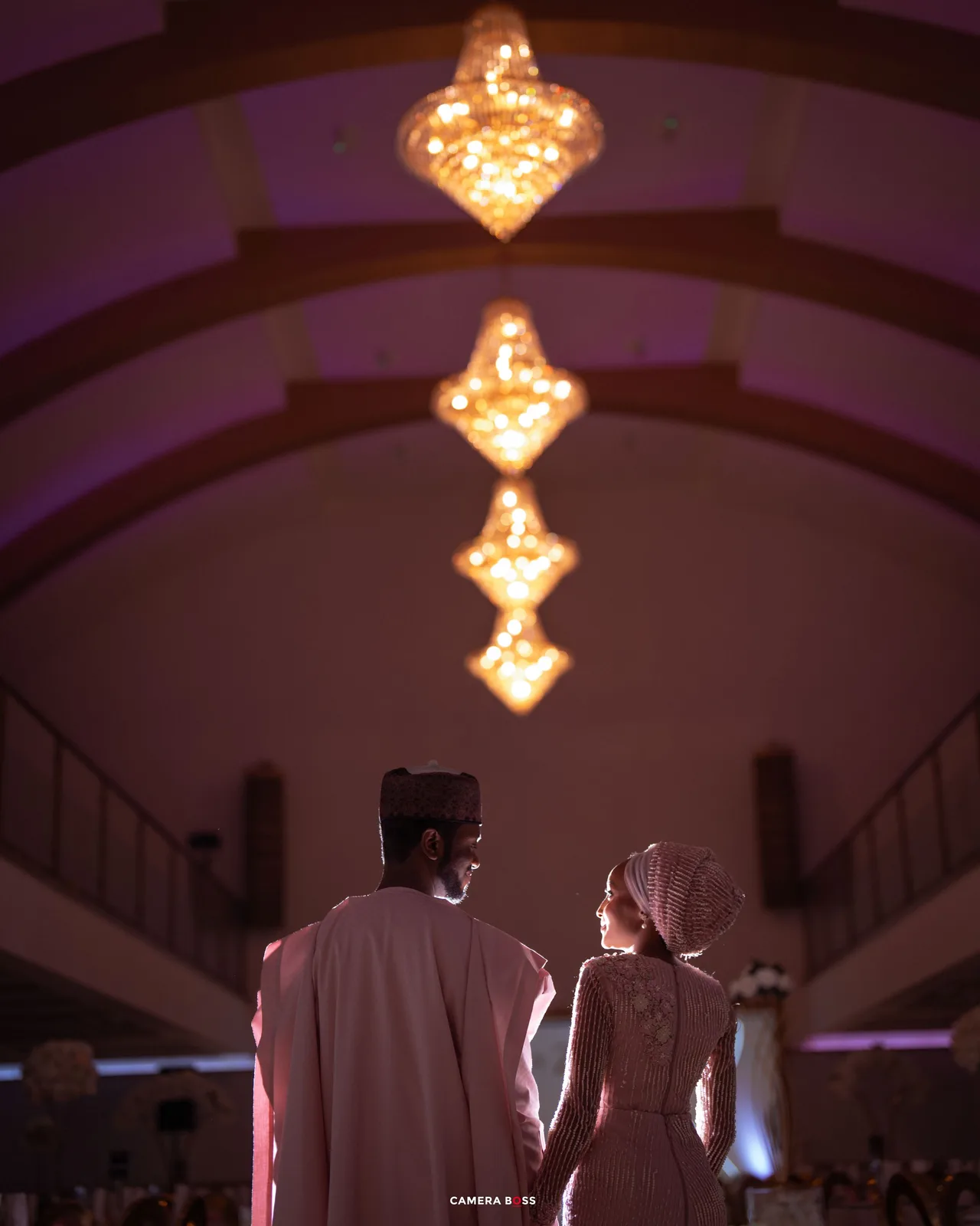 Elegant wedding couple holding hands under chandelier lights in a grand hall with purple ambient lighting.