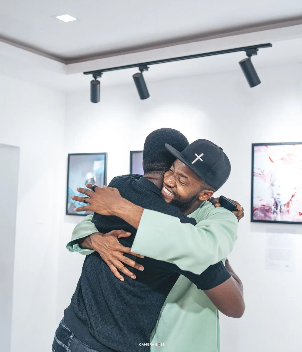 Two people share an emotional embrace in an indoor space with track lighting and framed artwork on the walls.