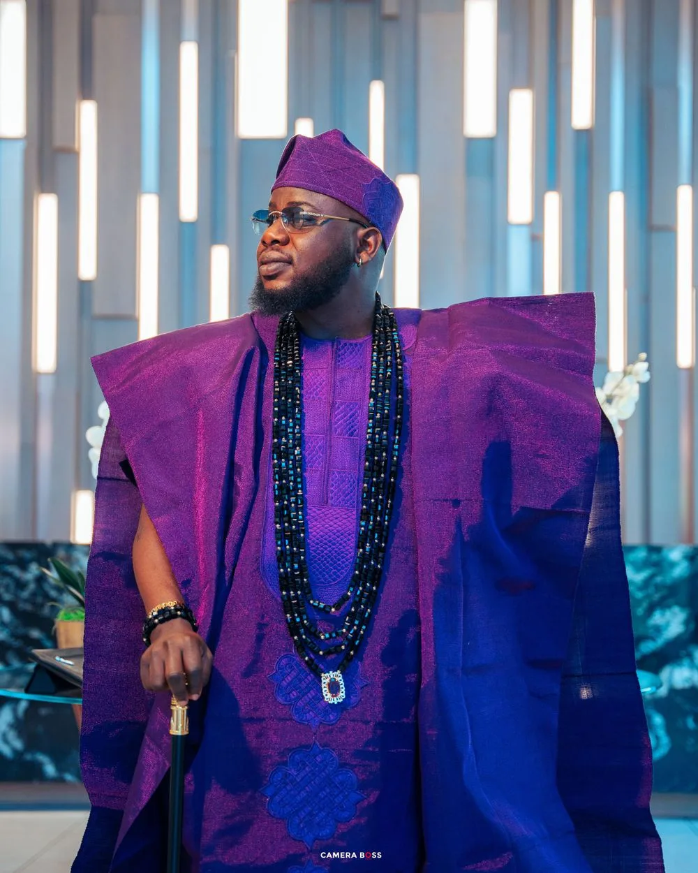 A person wearing an elegant purple and blue agbada with a beaded necklace stands against a modern illuminated backdrop.