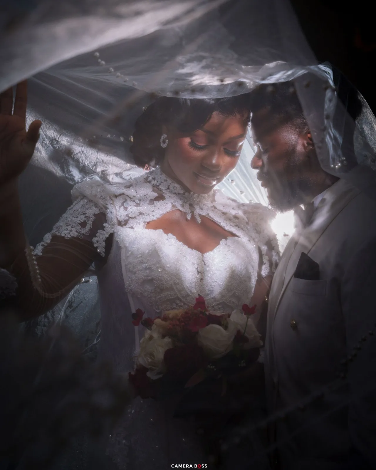 Bride and groom share an intimate moment under a white wedding veil, bride holding a bouquet of red and white flowers.