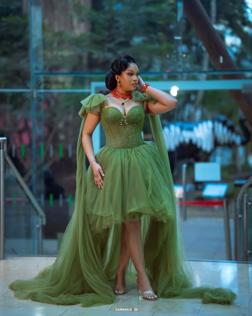 A person in an elegant olive green high-low tulle gown with flutter sleeves poses outdoors at dusk.