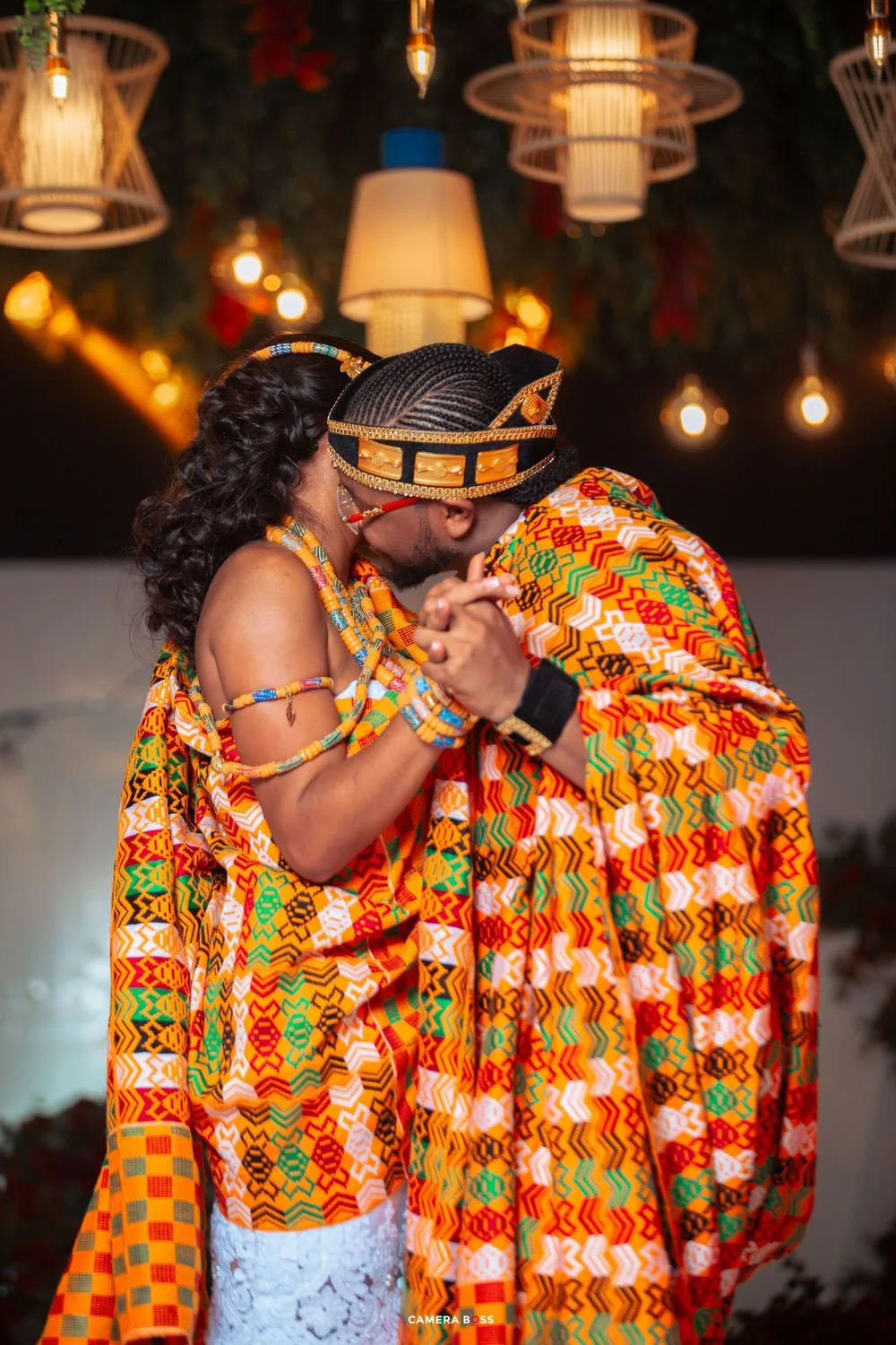 Couple in vibrant African kente cloth share an intimate moment under decorative hanging lights at an evening celebration.