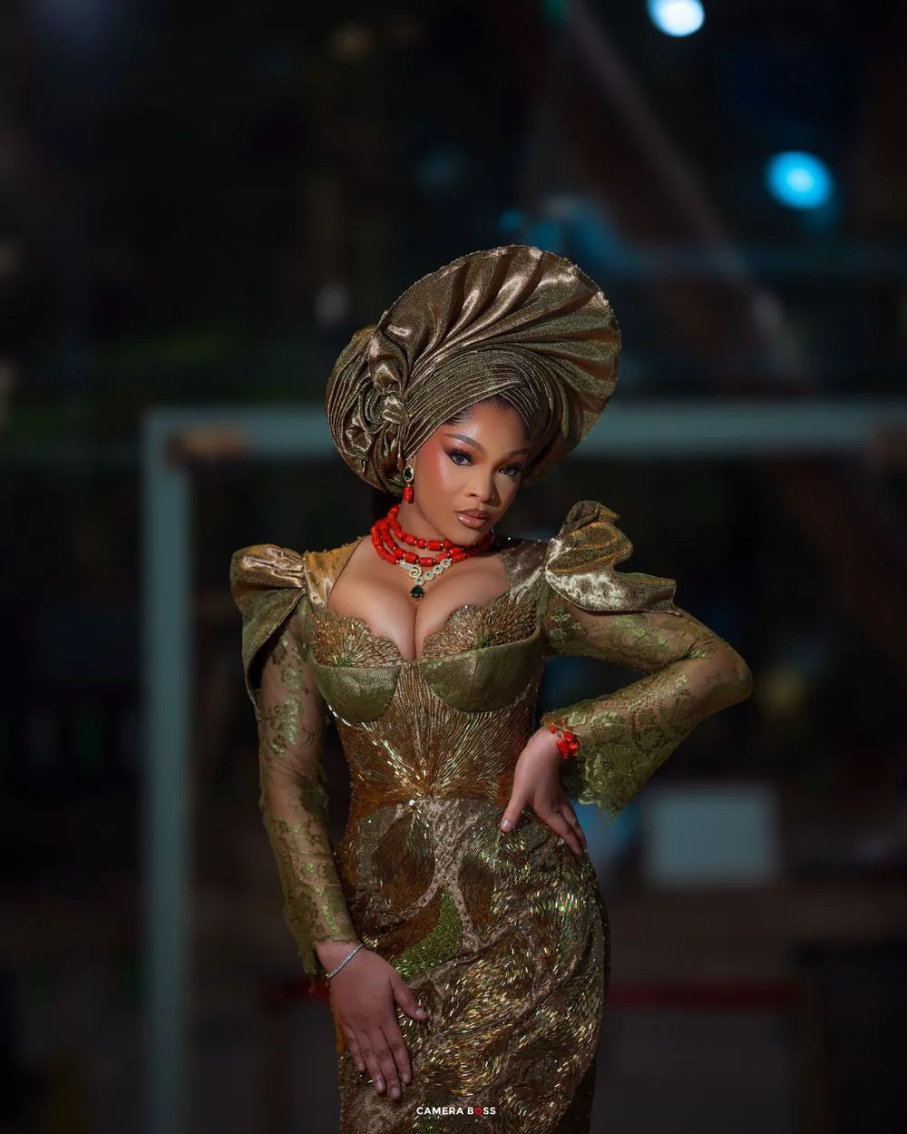 Elegant figure in a shimmering olive green dress with a dramatic sculptural headwrap and red statement necklace poses dramatically.