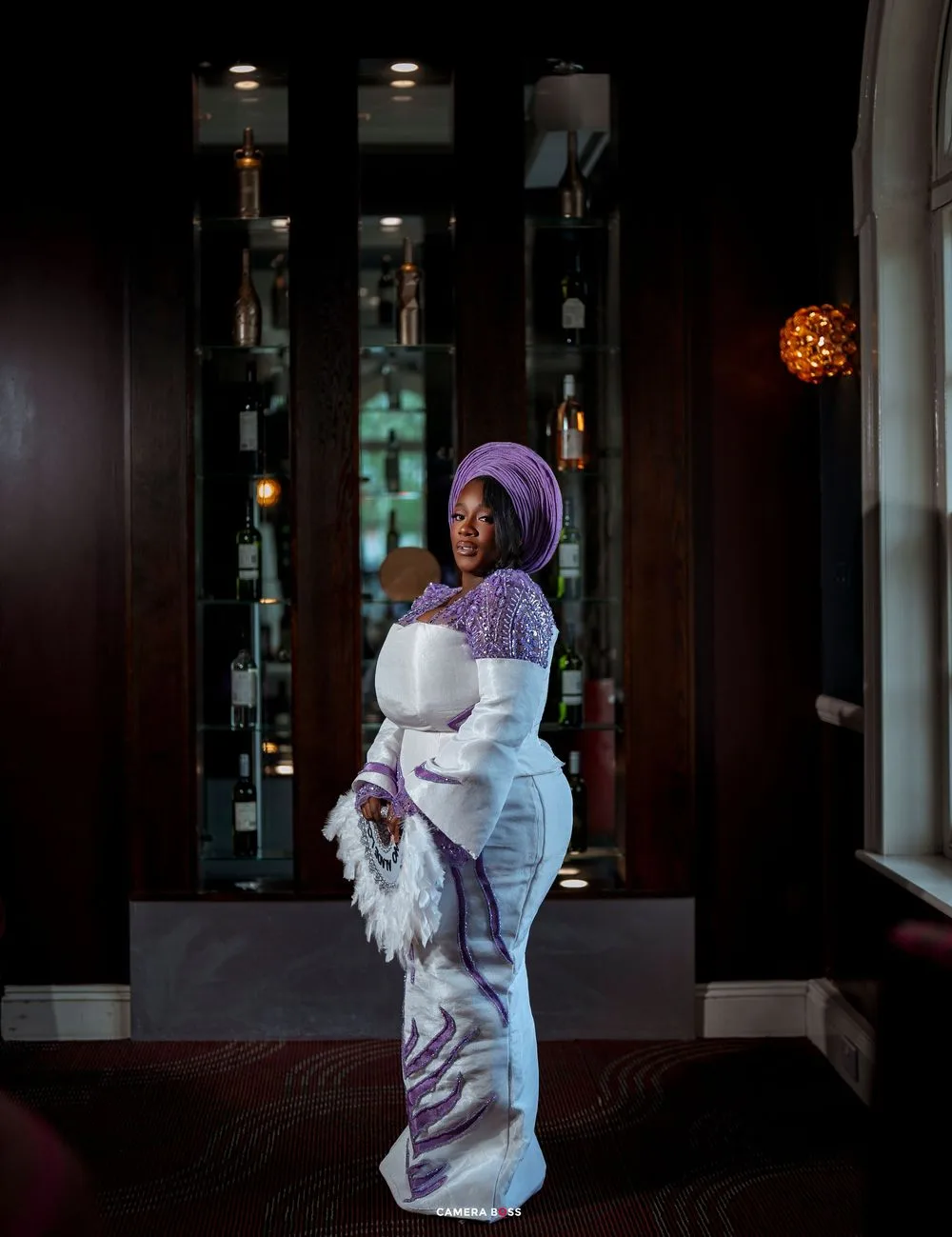 Woman in elegant white and purple traditional African attire with gele headwrap poses in a dimly lit bar.
