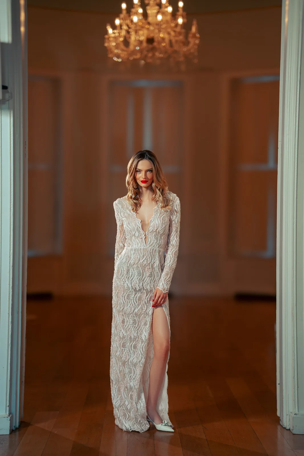 Elegant white lace wedding dress with plunging neckline and thigh-high slit under crystal chandelier in formal room.