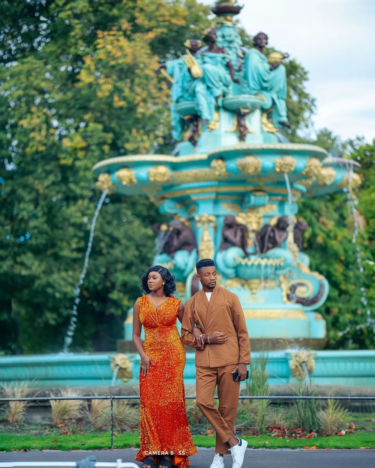 Well-dressed couple poses in front of ornate turquoise fountain with classical statues in a park setting.