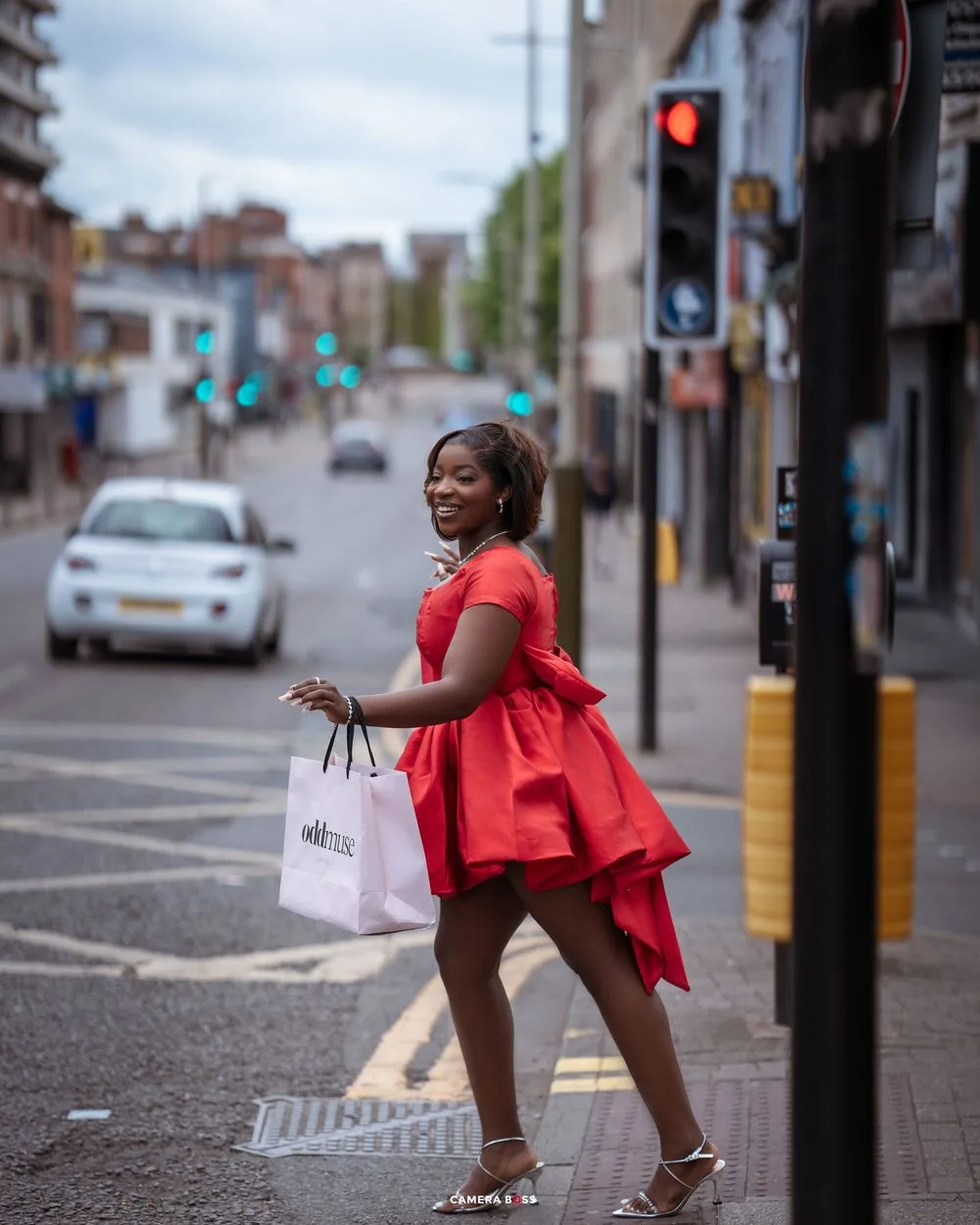 A woman in a flowing red dress carries a shopping bag while crossing an urban street on a cloudy day.