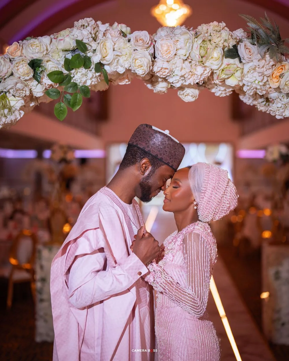 A romantic couple in matching pink traditional outfits embrace beneath a beautiful floral arch with white roses.