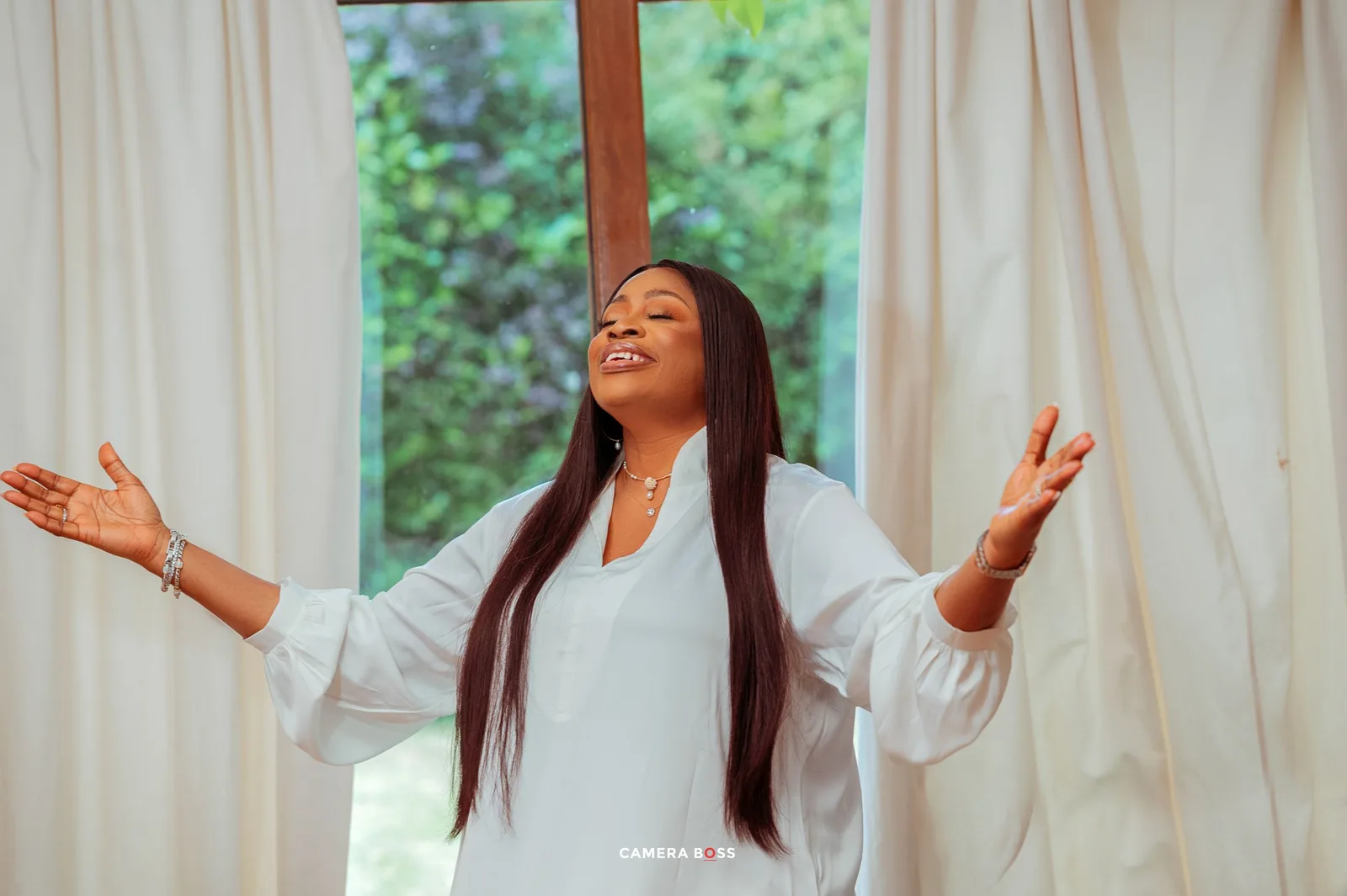 A person in white raises their arms in worship or celebration against a bright window backdrop.