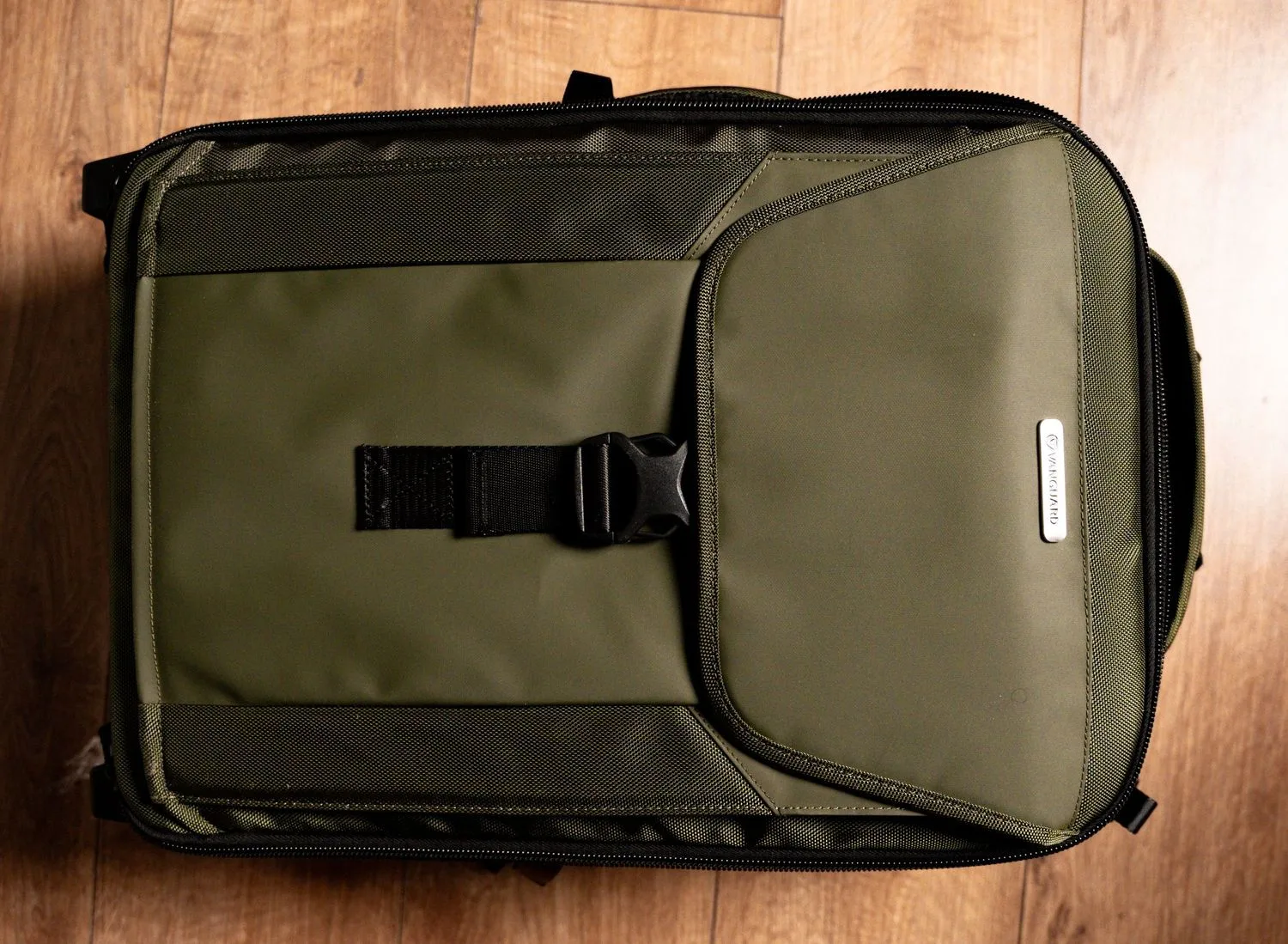 An olive green tactical backpack with black buckle strap and padded compartments sits on wooden surface.