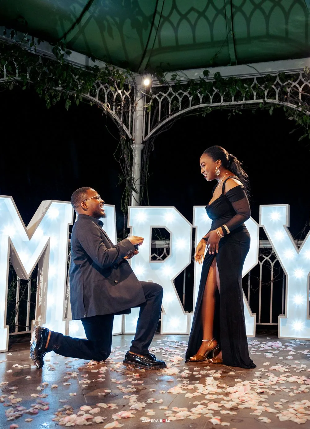 A romantic marriage proposal under a gazebo with lit up MARRY letters and rose petals scattered on the ground.
