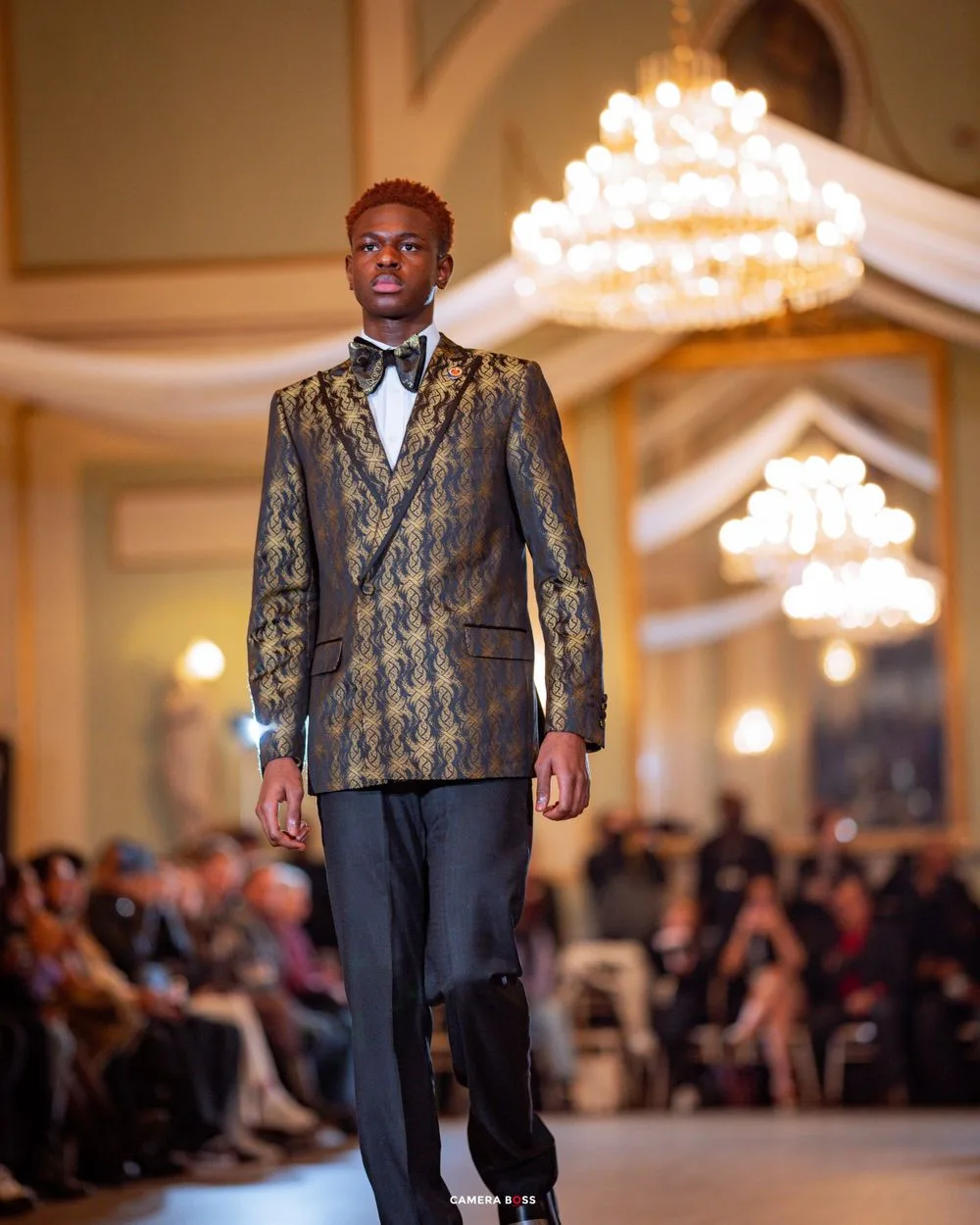 A model wearing a luxurious gold and black brocade double-breasted suit walks the runway under elegant crystal chandeliers.
