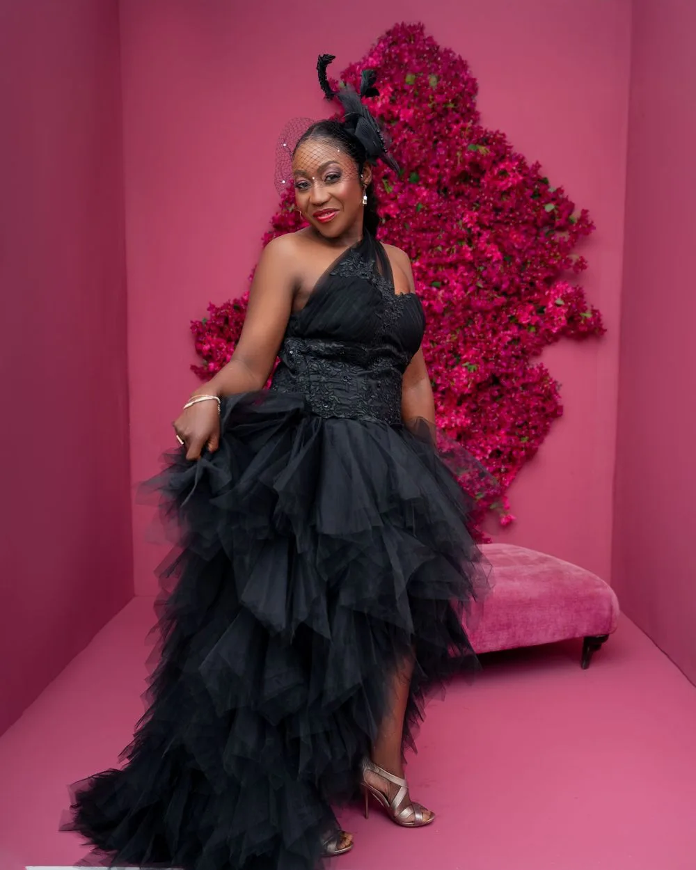 A dramatic black ruffled gown contrasts beautifully with vibrant pink floral backdrop in a pink-walled studio setting.
