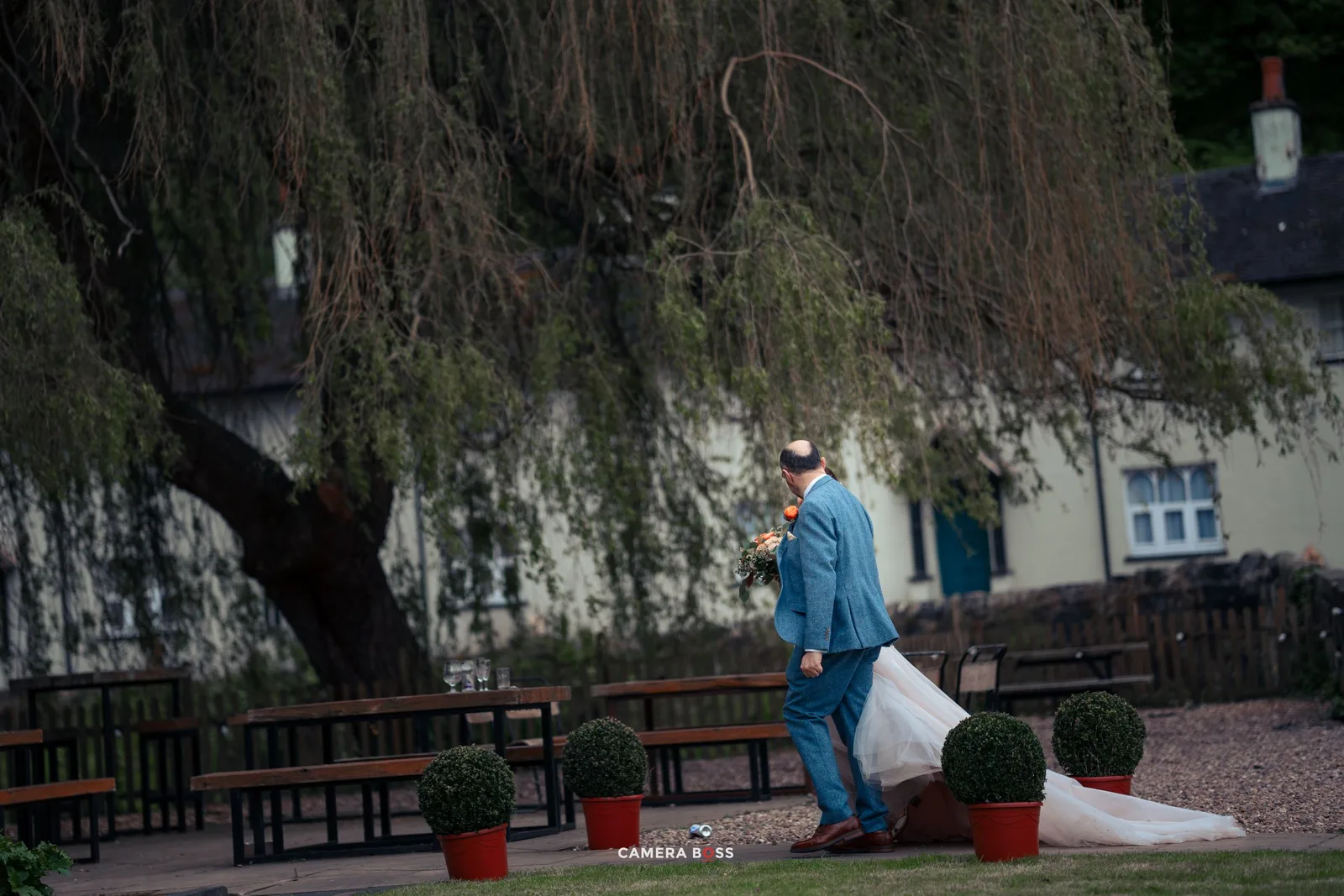 A person wearing protective gear walks with cleaning supplies through a courtyard with a weeping willow tree.