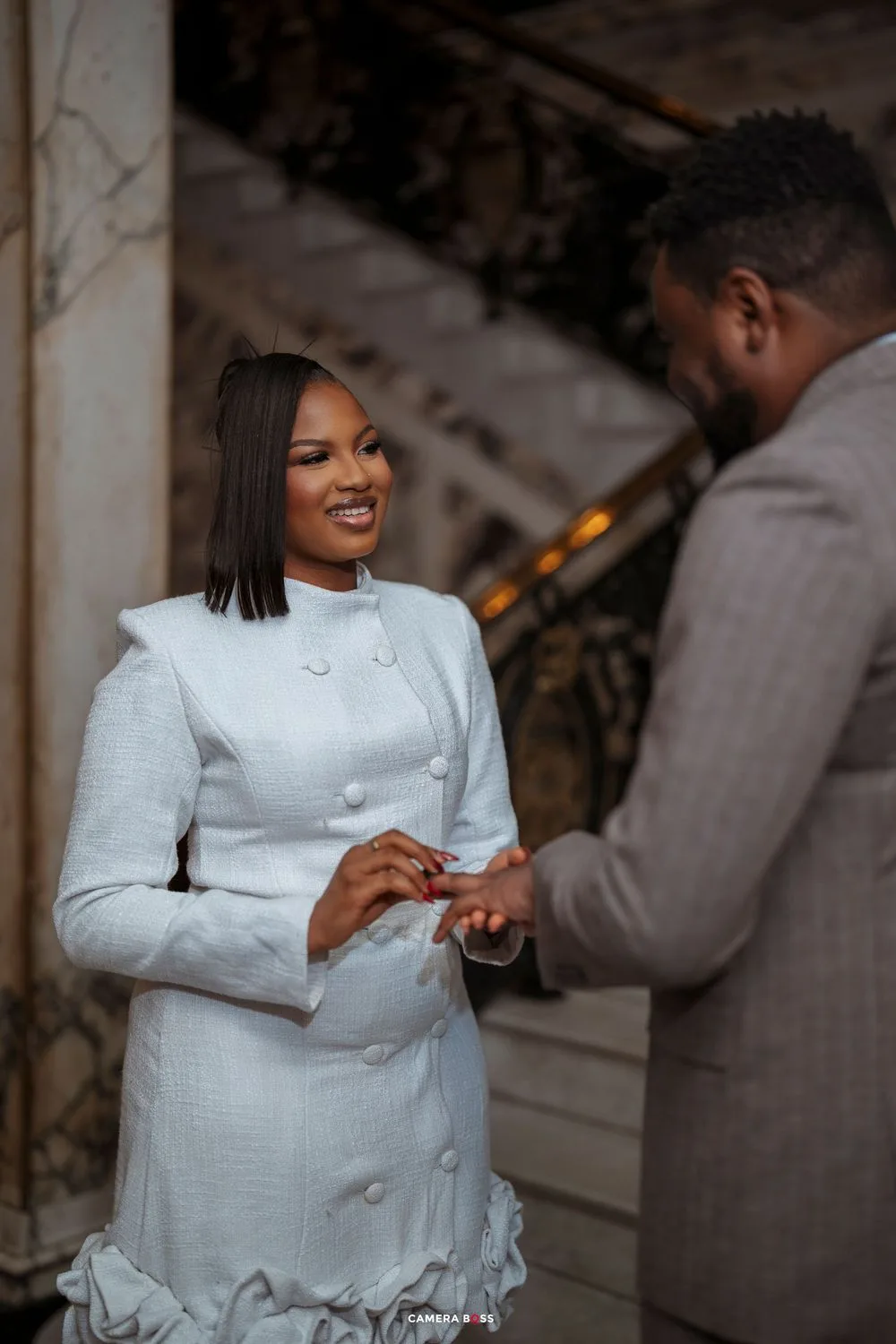 A smiling bride in a white double-breasted dress receives a ring from a groom in a gray suit during a wedding ceremony.
