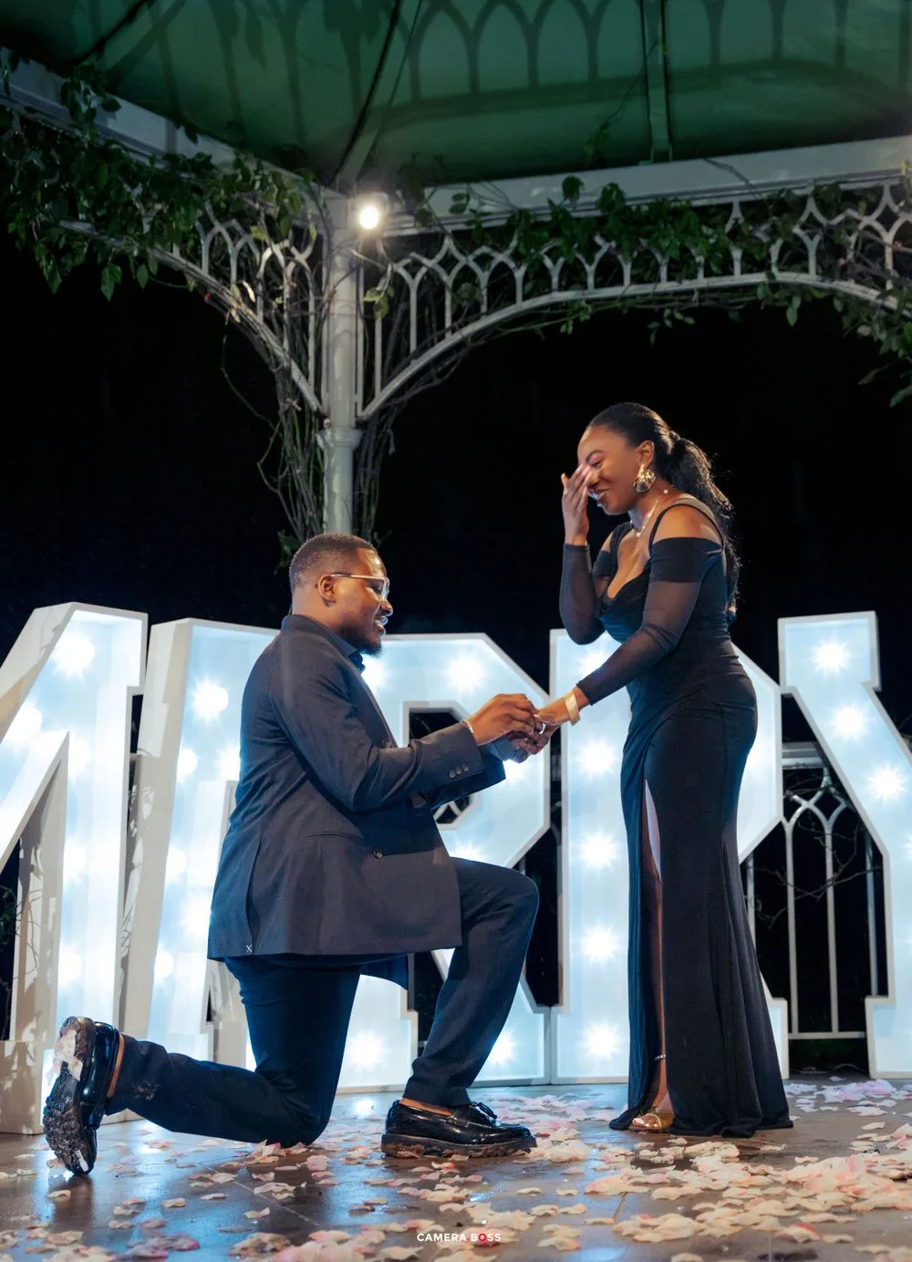 A romantic proposal moment under an ornate gazebo at night with illuminated letters in the background.