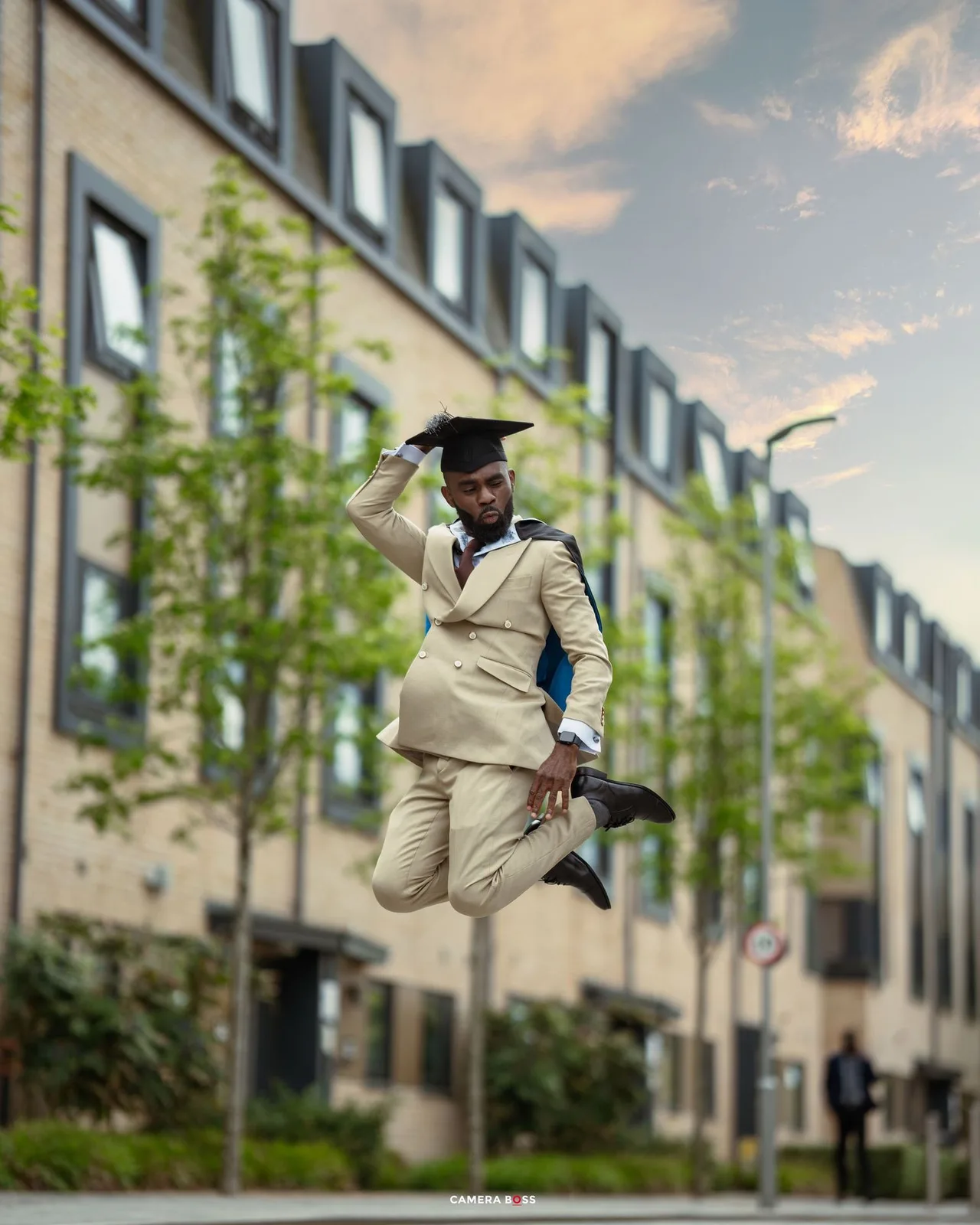 Graduate in a light colored suit and cap jumps joyfully in front of a modern apartment building on graduation day.