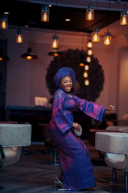 Dancing figure in flowing purple and blue traditional dress with head wrap stands in restaurant with hanging lanterns.