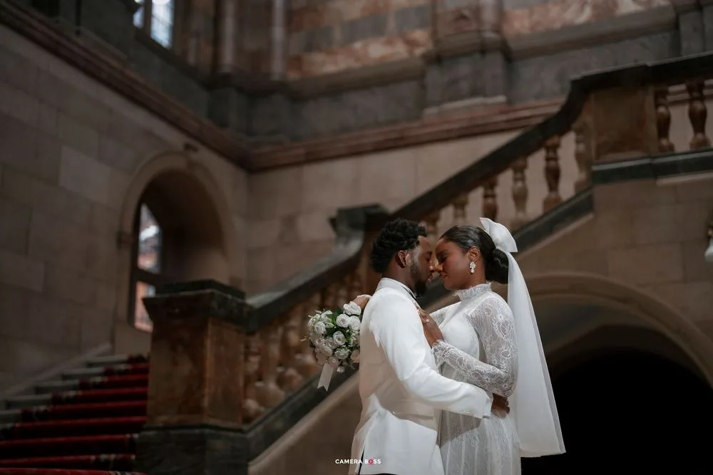 Two brides in white gowns share an intimate moment on grand staircase holding bouquet.