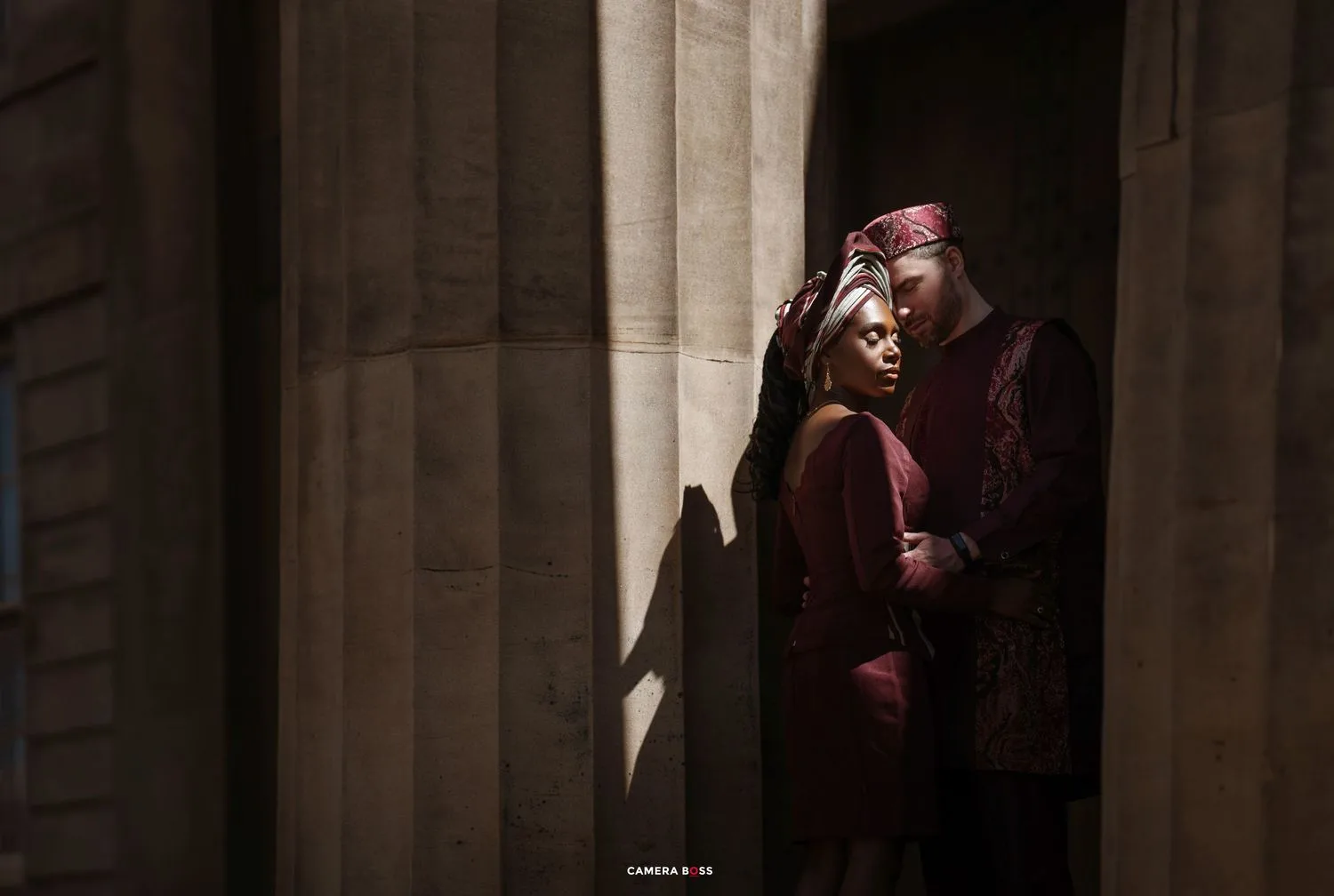 A person in maroon clothing stands in shadows between stone columns with dramatic lighting.