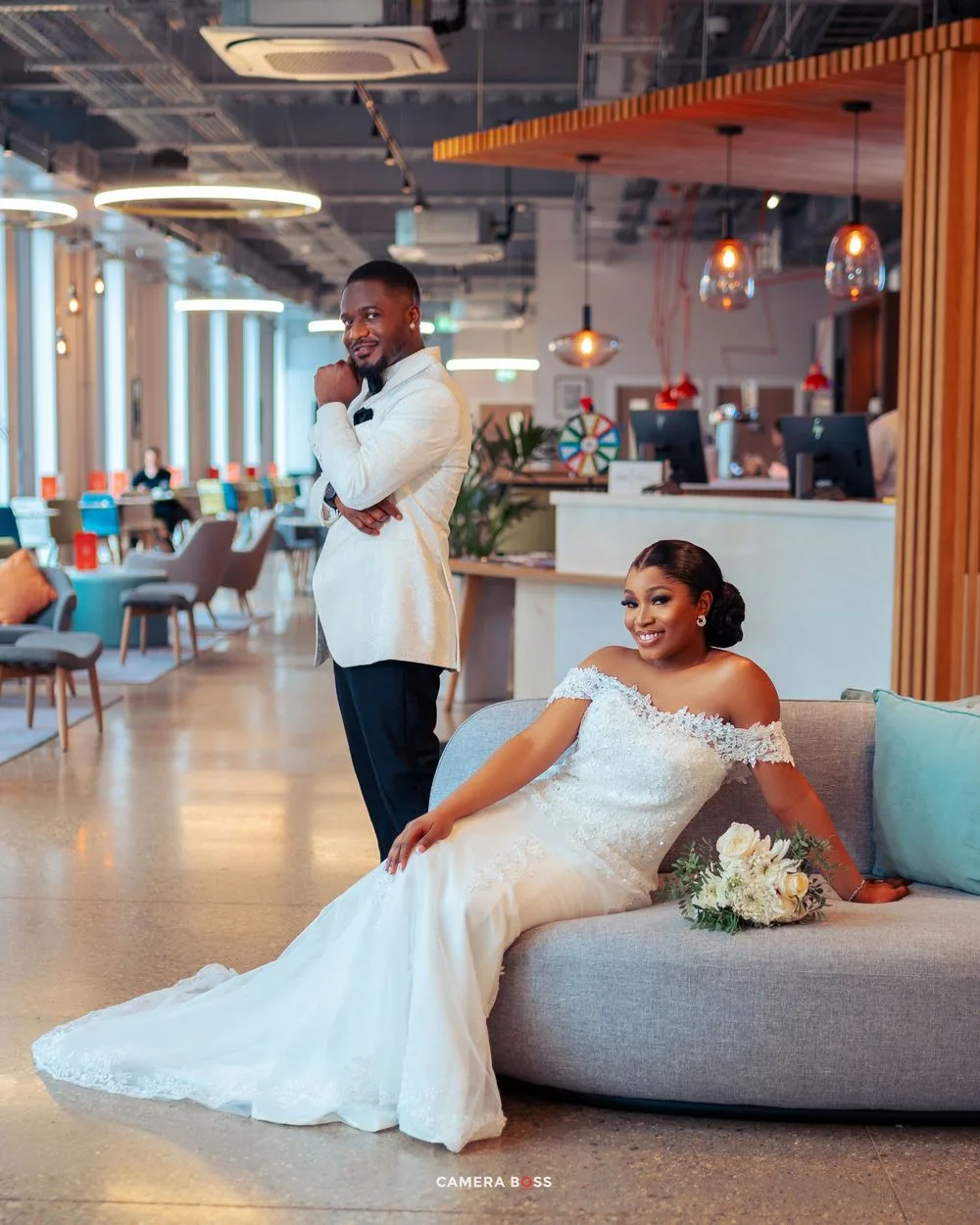 Elegant bride in off-shoulder white gown relaxes on gray couch while groom stands behind in modern dining space.