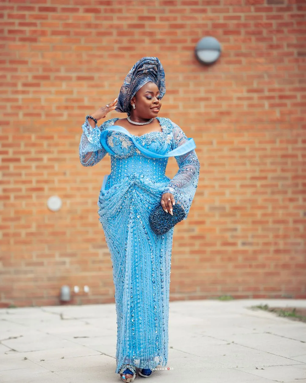 Elegant turquoise blue lace gown with long bell sleeves and sequin details stands out against rustic brick wall backdrop.