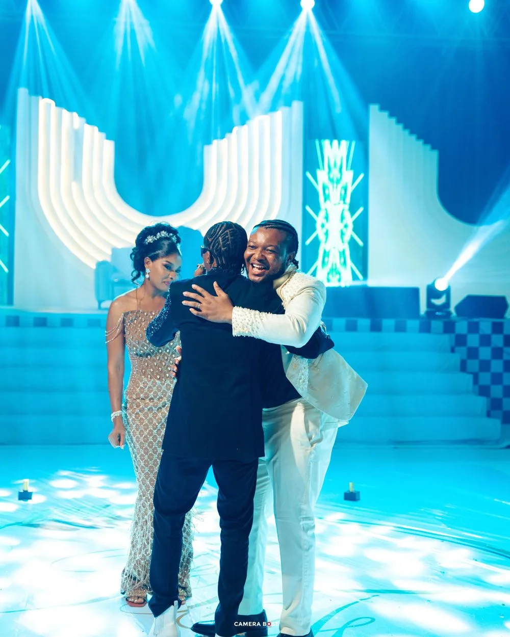 People embracing on an elegant stage with bright blue lighting and white geometric set pieces in the background.