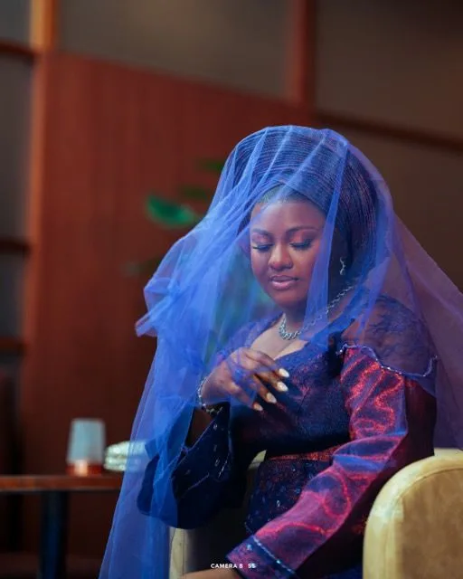 Woman wearing a blue veil and red patterned dress sits while looking down with a gentle smile.