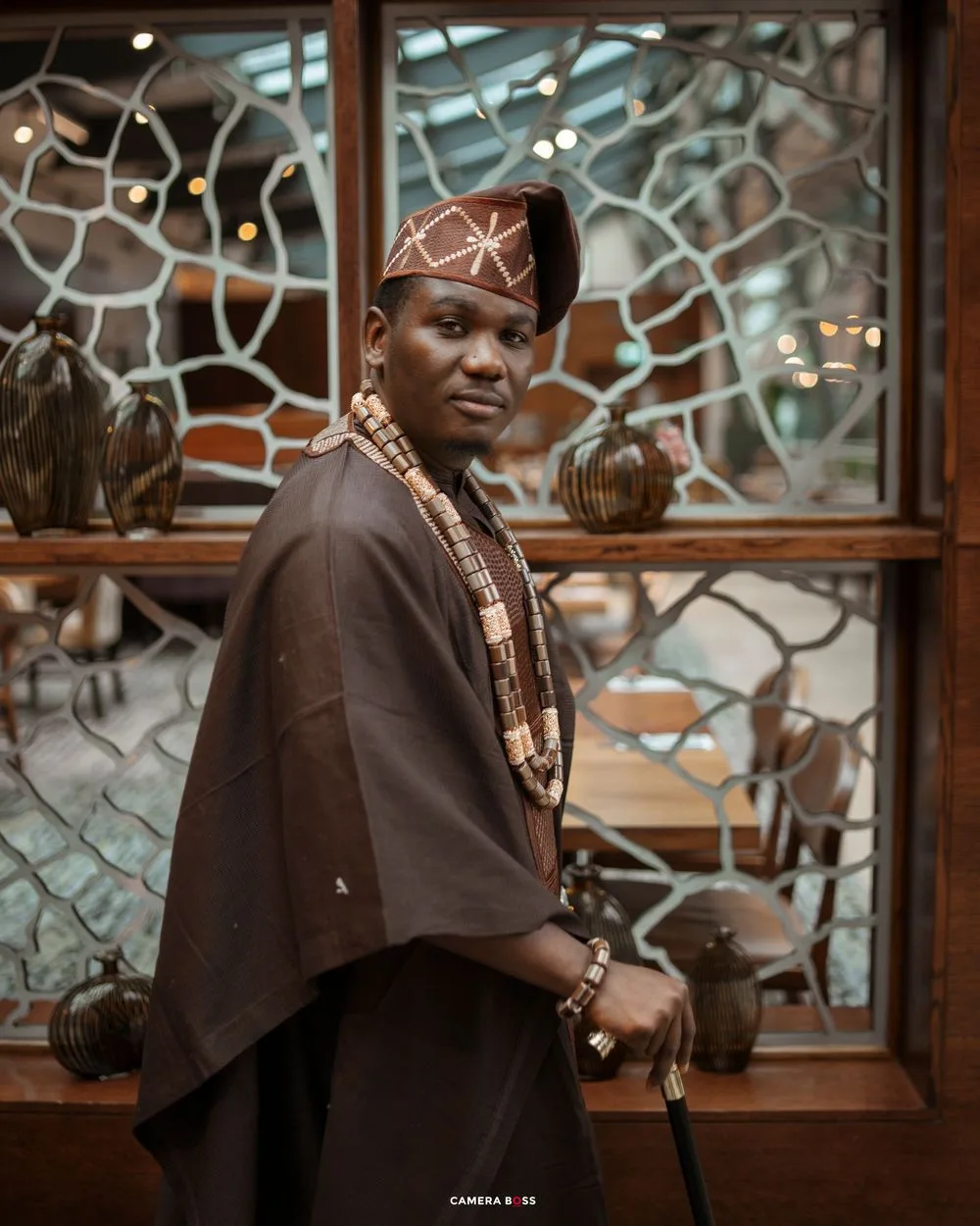 Man in traditional African attire with beaded necklace, cap, and walking stick poses before decorative wooden window.
