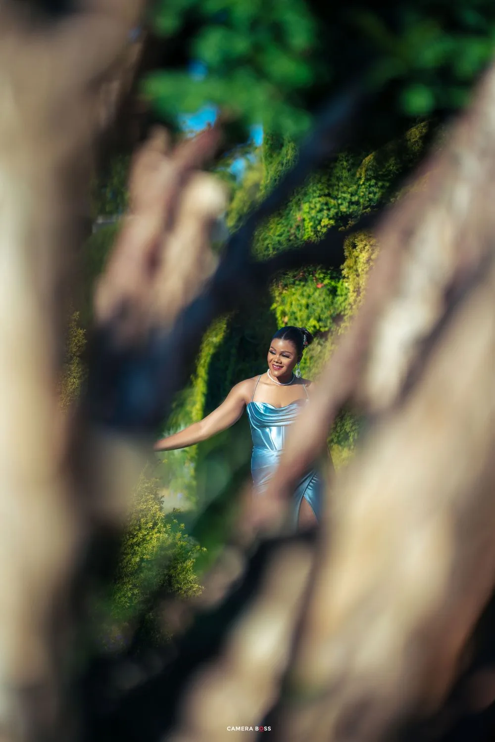 A dancer in a blue dress performs gracefully in a garden setting framed by blurred tree trunks and green foliage.