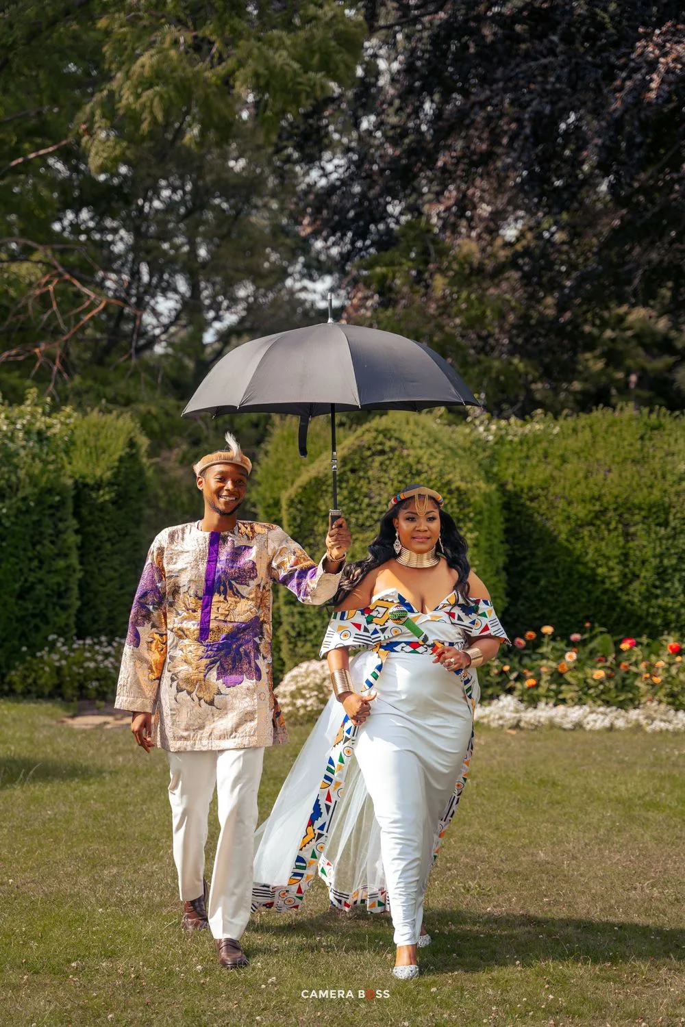 Couple walking in a garden under a grey umbrella wearing colorful traditional ethnic clothing on a sunny day.