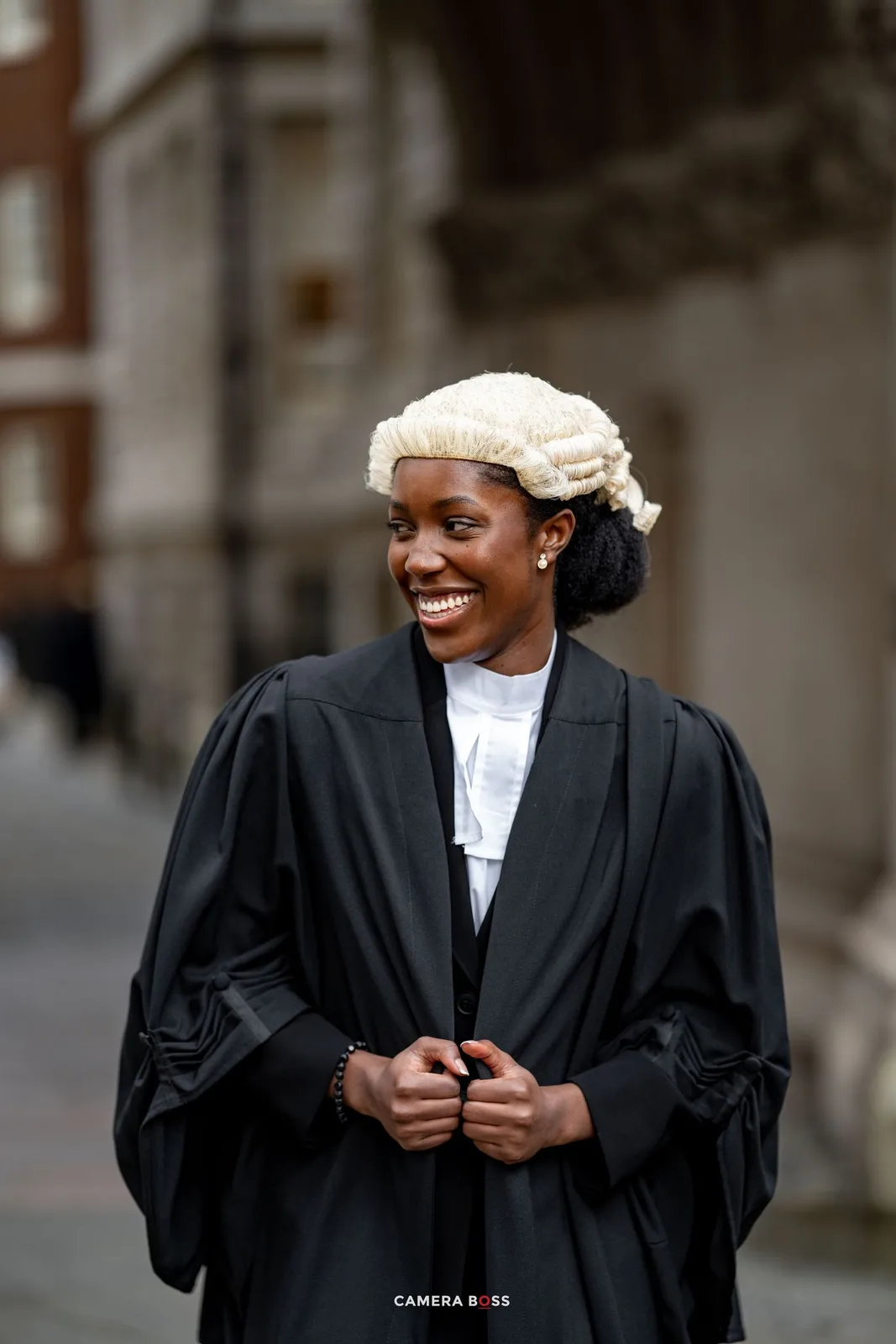 A legal professional in traditional black court robes and white barrister's wig smiles while standing outside a building.