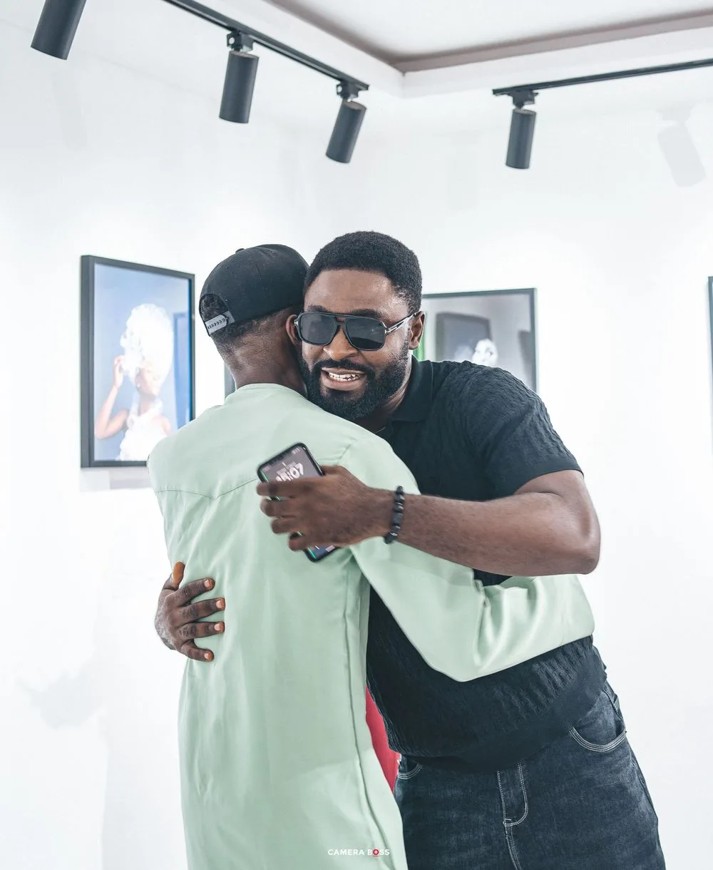 Two people share a warm embrace in an art gallery with track lighting and framed artwork on the walls.