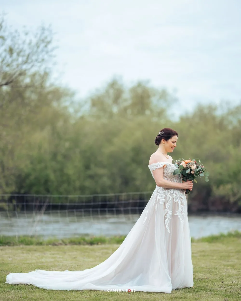 A bride wearing an elegant off-shoulder white wedding gown with a flowing train stands beside a lake holding a floral bouquet.