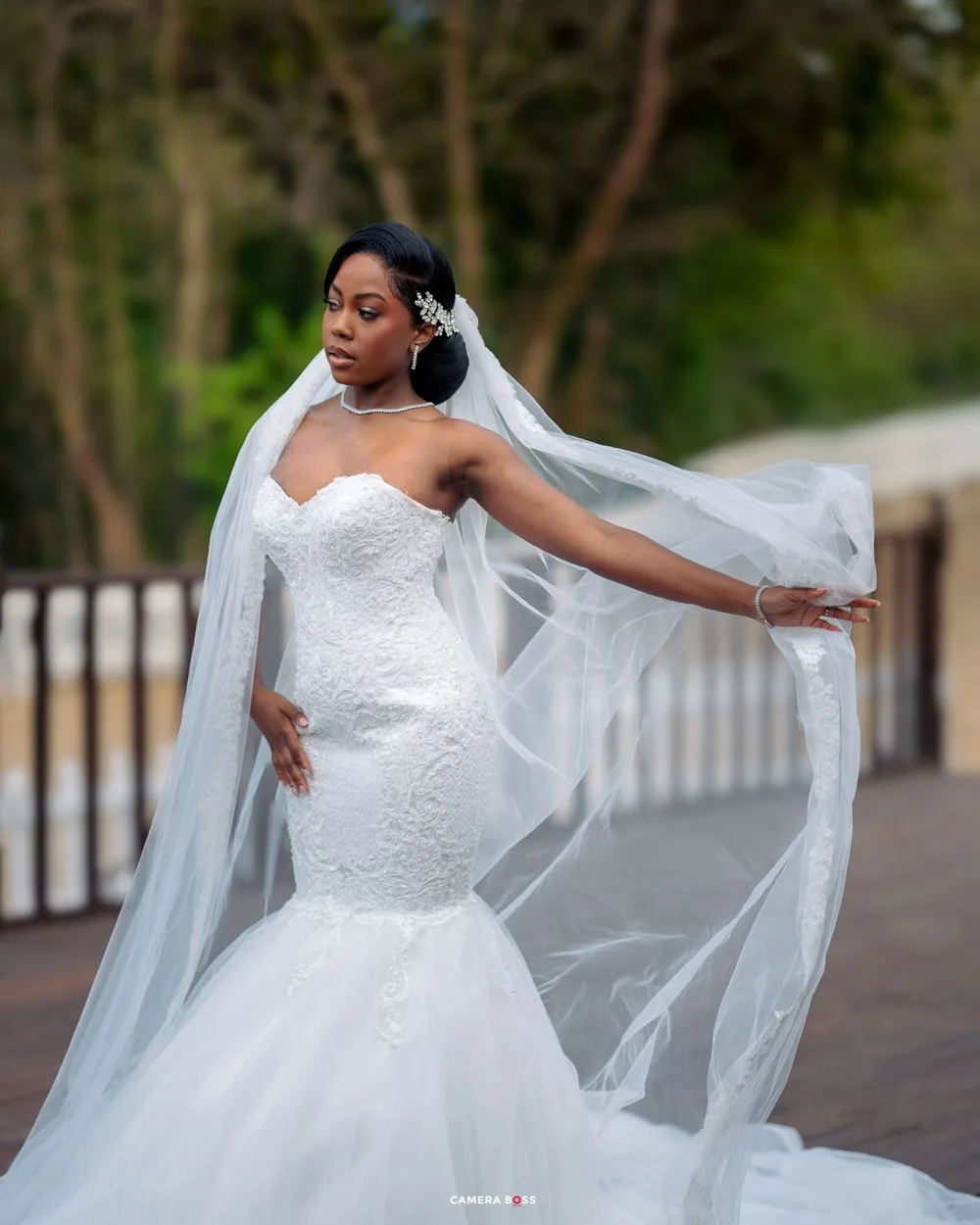 A bride in a lace mermaid wedding gown poses elegantly with her flowing veil on an outdoor walkway.