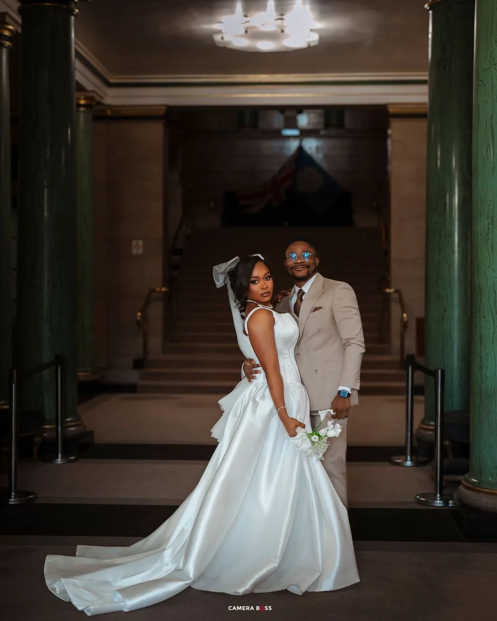 A couple poses elegantly in formal wedding attire inside an ornate hallway with green columns and chandelier lighting.
