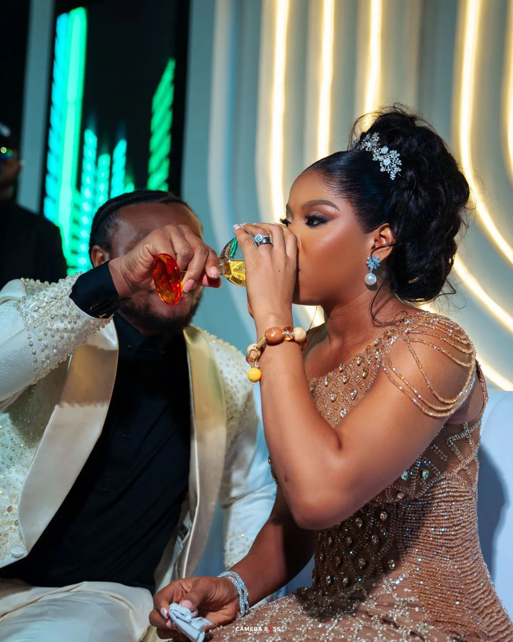A couple takes a celebratory toast together at their wedding reception, dressed in formal white and beige attire.