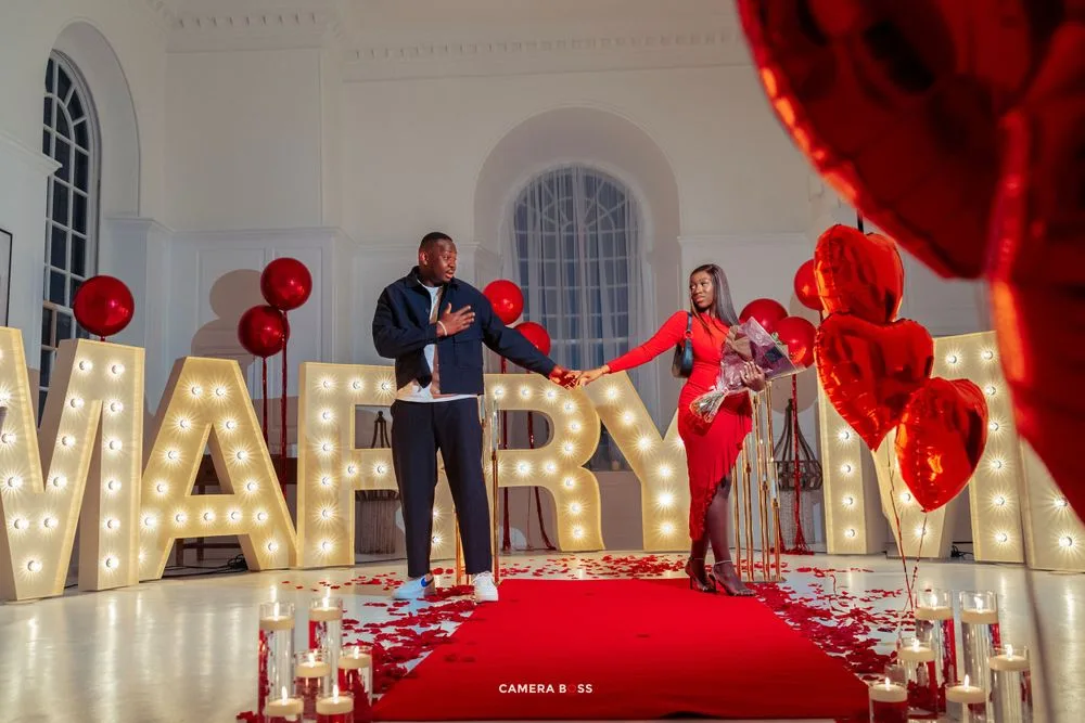 A romantic proposal setup with illuminated MARRY ME letters, red balloons, and a couple standing on a red carpet.