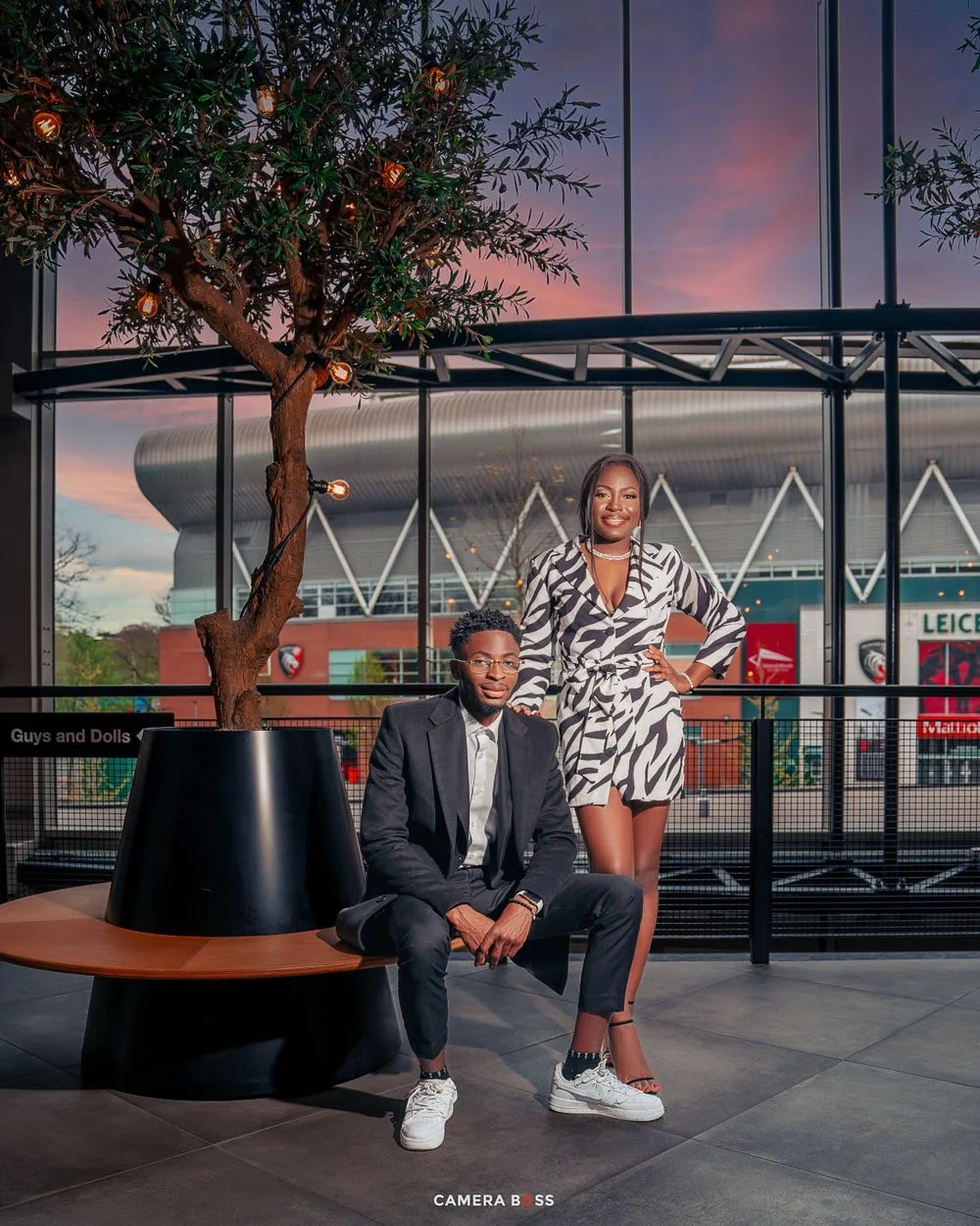 A stylish couple poses at dusk outside a modern building with geometric architecture and a pink sunset sky.