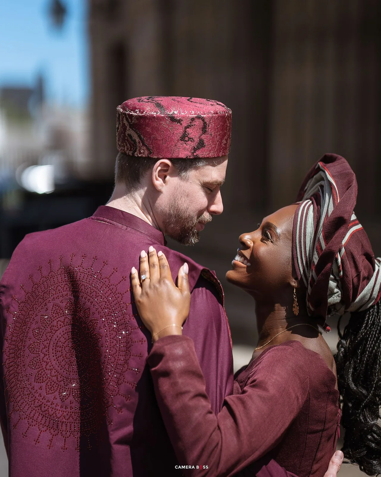 A couple in burgundy colored outfits shares a tender romantic moment while smiling at each other outdoors.