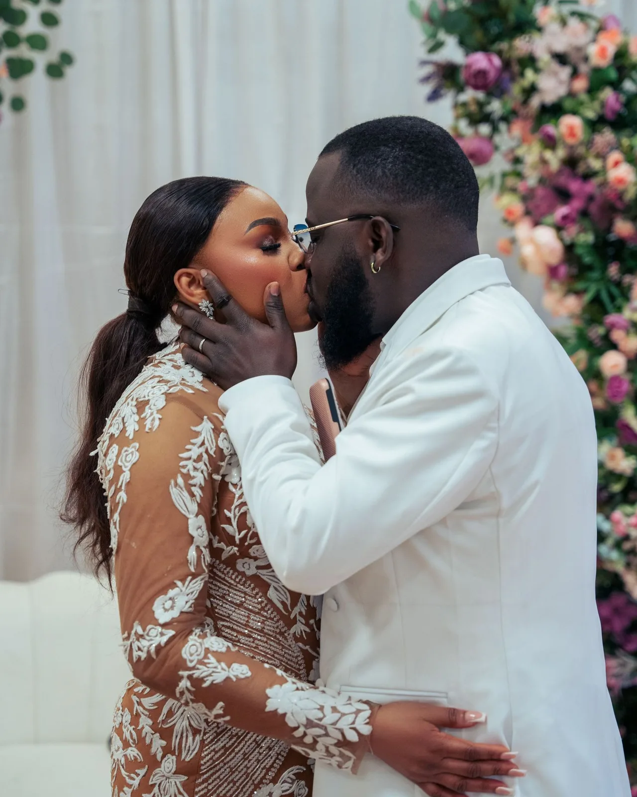 A loving couple shares a romantic kiss in front of a floral backdrop at a wedding celebration.