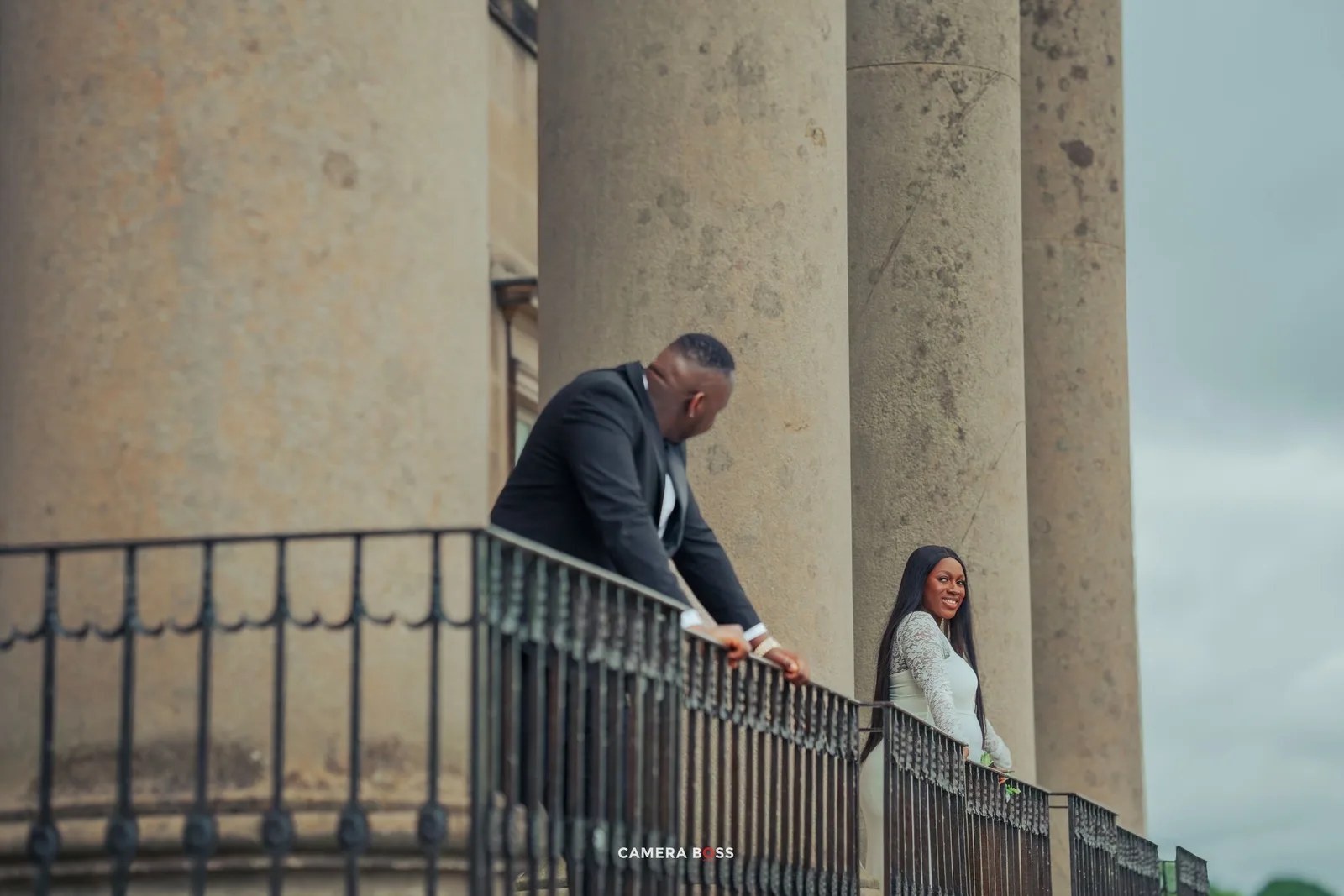 Two people stand against a railing near tall stone columns on a cloudy day.